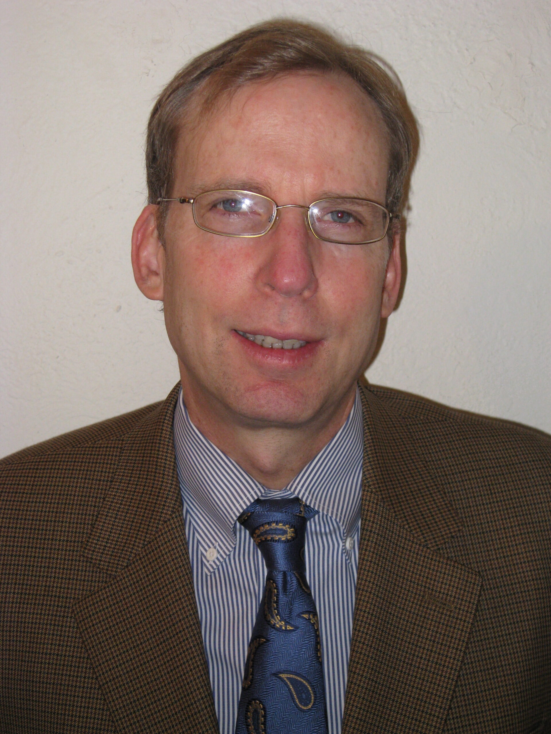 A man wearing a blue tie, and brown suit jacket with glasses smiling.